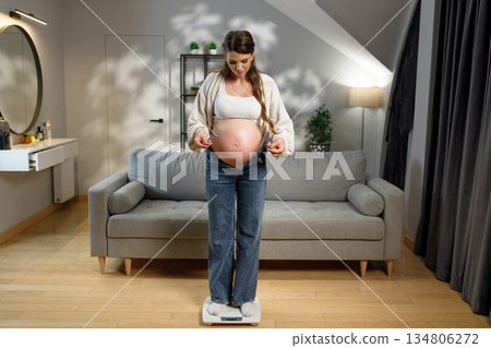 Expecting mother stands on scale in living room, checking weight gain during pregnancy, feeling clothes tightness Expecting mother stands on scale in living room, checking weight gain during pregnancy, feeling clothes tightness 134806272