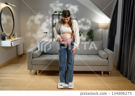 Pregnant woman stands on scale measuring her abdomen with tape measure in living room Pregnant woman stands on scale measuring her abdomen with tape measure in living room 134806275