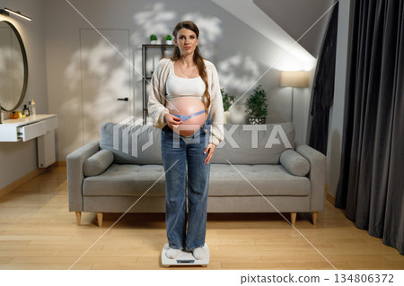 Pregnant woman stands on scale measuring her belly circumference with tape measure in living room 134806372