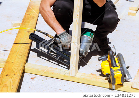 Carpenter works on construction site using nail gun to attach wooden beams pieces together Carpenter works on construction site using nail gun to attach wooden beams pieces together 134811275