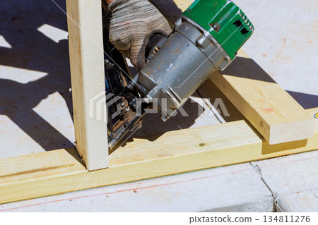 Carpenter holds tool to pneumatic nail gun wooden pieces while working on construction site 134811276