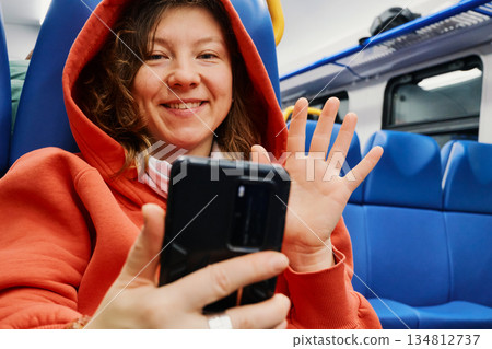 Smiling woman waving at smartphone on train, enjoying connectivity during travel Smiling woman waving at smartphone on train, enjoying connectivity during travel 134812737