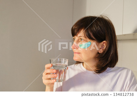 Mature Woman applying under eye patches and drinking water for morning self-care and wellness Mature Woman applying under eye patches and drinking water for morning self-care and wellness 134812738