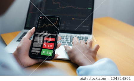 Investor Using Smartphone and Laptop for Cryptocurrency Trading and Stock Market Analysis 134814801