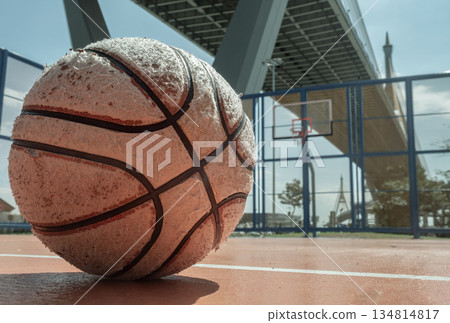 A heavily used basketball rests on an outdoor court, with a hoop and fiberglass backboard visible beneath the industrial Ring Road Bridge in the background. 134814817