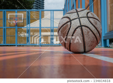 A weathered basketball rests on the vibrant orange outdoor court, with the hoop and fiberglass backboard visible in the background. 134814818