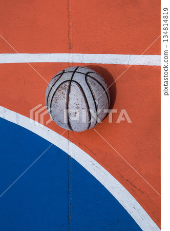 A weathered basketball sits in stark contrast against the vibrant orange and blue expanse of the court, defined by crisp white lines. 134814819