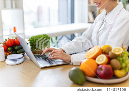 A nutritionist smiles while working on a laptop surrounded by fresh fruits and vegetables, promoting healthy eating habits. 134817106