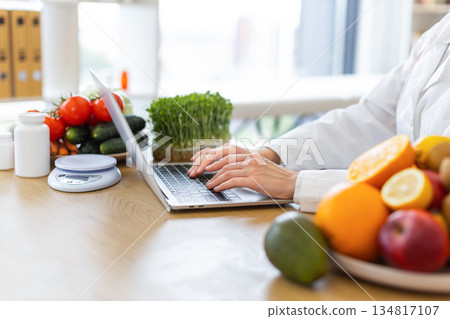 A nutritionist is working on a laptop surrounded by fresh fruits, vegetables, and supplements, emphasizing healthy eating. 134817107