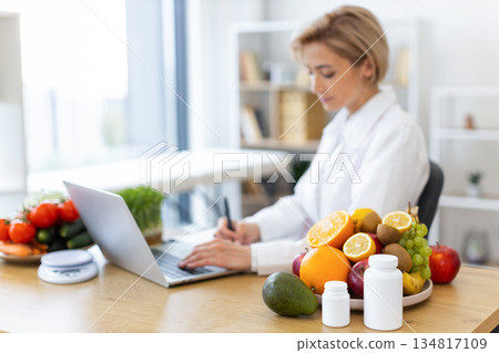 A nutritionist is working on a laptop surrounded by fresh fruits and vegetables, promoting healthy eating and wellness. 134817109