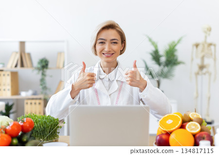 A smiling dietitian gives a thumbs up while working on her laptop, surrounded by healthy food. 134817115