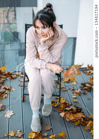Woman sits in meditative pose on balcony with autumn leaves. Hygge lifestyle, mental health awareness, seasonal affective disorder. 134817194