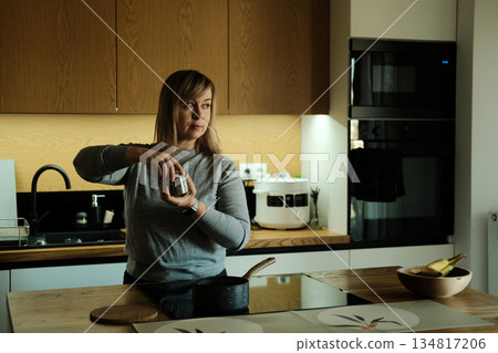 Woman stands in modern kitchen holding jar of herbs while looking away from camera. Natural flavor enhancers, plant-based nutrition, holistic cooking, wellness kitchen. 134817206
