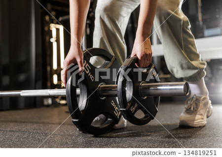 Close up of female athlete adding weight plates to barbell for strength workout in modern fitness gym Close up of female athlete adding weight plates to barbell for strength workout in modern fitness gym 134819251