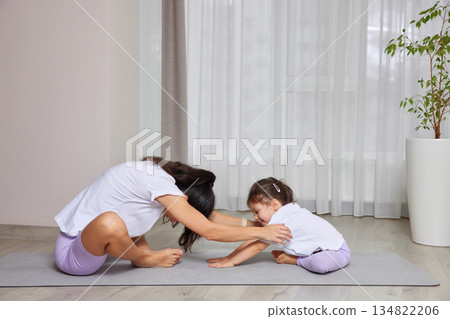 Mother helping young daughter stretching on a yoga mat during home workout 134822206