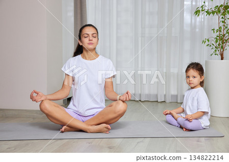 Mother and daughter sitting in lotus pose on yoga mats at home 134822214
