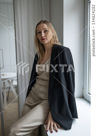 Elegant woman in beige dress and black blazer sitting by window in minimalist interior 134824282