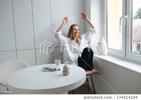 Relaxed woman in white shirt sitting by window in bright minimalist home interior 134824284