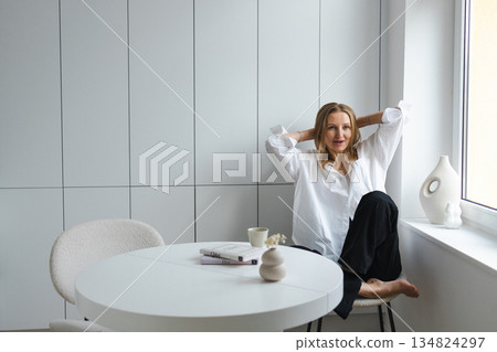 Relaxed woman in white shirt sitting by window in bright minimalist home interior Relaxed woman in white shirt sitting by window in bright minimalist home interior 134824297