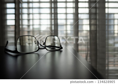 Eyeglasses on the table and light filtering through the curtains from the large window. Starting the day in the morning. Eyeglasses on the table and light filtering through the curtains from the large window. Starting the day in the morning. 134828905