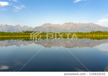Clear reflection of green forest and rugged mountains on calm lake surface Clear reflection of green forest and rugged mountains on calm lake surface 134829242