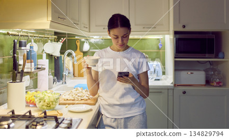 Woman checking smartphone while holding cup in cozy home kitchen. Woman checking smartphone while holding cup in cozy home kitchen. 134829794