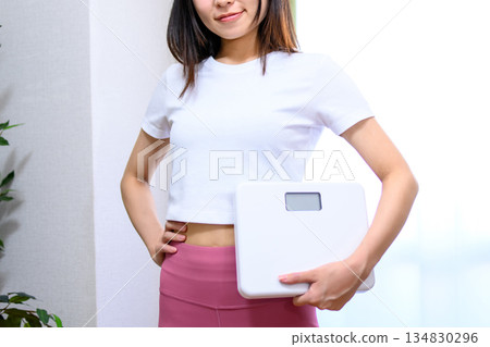 一位微笑的日本女性身穿瑜珈服，手持體重秤，穿著緊身運動褲，臉部模糊，照片為局部放大。 134830296