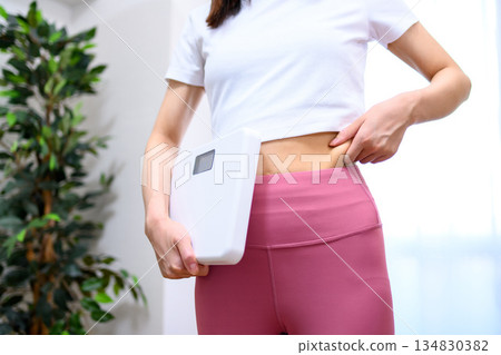 Japanese woman in yoga wear holding a scale and pinching her belly fat. No face. Diet. Leggings. Japanese woman in yoga wear holding a scale and pinching her belly fat. No face. Diet. Leggings. 134830382