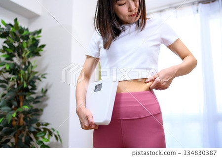 Japanese woman in yoga wear holding a scale and pinching her belly fat. No face. Diet. Leggings. 134830387
