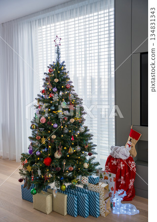 Plastic Christmas tree with colorful ornaments and wrapped gifts in bright living room Plastic Christmas tree with colorful ornaments and wrapped gifts in bright living room 134831343