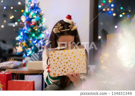 Woman with reindeer headband looking into Christmas gift box near decorated tree with lights indoor Woman with reindeer headband looking into Christmas gift box near decorated tree with lights indoor 134831349