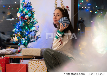 Young woman with reindeer headband holding small wrapped gift near glowing tree at home Young woman with reindeer headband holding small wrapped gift near glowing tree at home 134831350