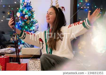 Young woman with long hair making a video call near Christmas tree and gifts in cozy festive room. Young woman with long hair making a video call near Christmas tree and gifts in cozy festive room. 134831351