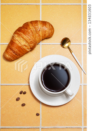 A cup of freshly brewed black coffee and a croissant with coffee beans and spoon on orange tiles. 134831603