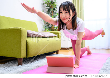 獵犬:一位年輕女子在家中一邊用平板電腦觀看 YouTube 視頻,一邊做瑜伽。身穿瑜珈服。 獵犬:一位年輕女子在家中一邊用平板電腦觀看 YouTube 視頻,一邊做瑜伽。身穿瑜珈服。 134832683