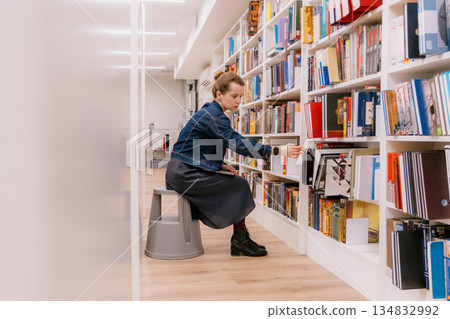 woman is sitting at shelves in library, student is choosing book. library is filled with books. woman is sitting at shelves in library, student is choosing book. library is filled with books. 134832992