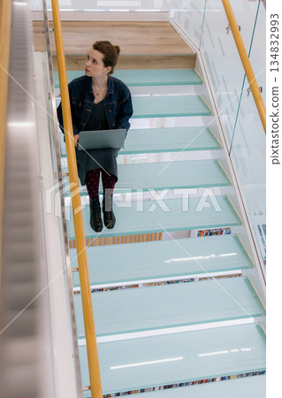 young woman is sitting on glass staircase with laptop in front of her and working. business person. young woman is sitting on glass staircase with laptop in front of her and working. business person. 134832993