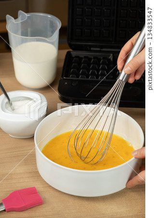 Hand Using a Whisk to Mix Raw Eggs in a White Bowl for Baking Waffles 134833547