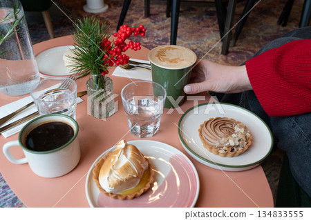 Cafe table with two plated desserts, latte in green cup, glass of water, and person in red sweater reaching for coffee, creating cozy indoor cafe mood 134833555
