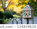 A charming, hand-painted birdhouse sits atop a white picket fence, in a picturesque autumnal garden setting 134834628