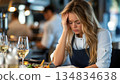 A young chef, overwhelmed and stressed, sits at a restaurant table, hand on her forehead, showing exhaustion and worry 134834638