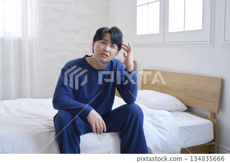 A man in his 20s sitting on the bed with a headache A man in his 20s sitting on the bed with a headache 134835666