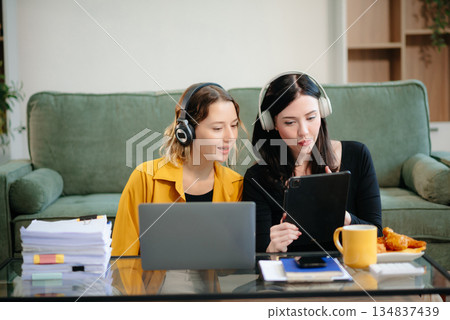 Two women collaborating on a project at home, brainstorming with a laptop and notebook in a cozy setting. Two women collaborating on a project at home, brainstorming with a laptop and notebook in a cozy setting. 134837439