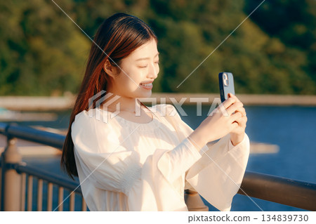 A Japanese woman looking at her smartphone on a bank with the sea in the background A Japanese woman looking at her smartphone on a bank with the sea in the background 134839730