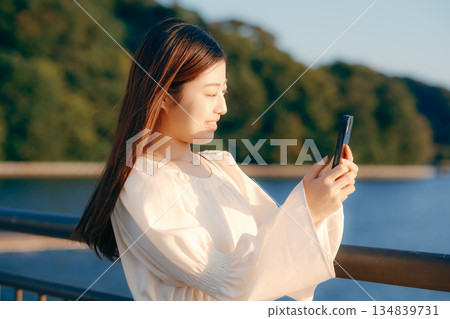 A Japanese woman looking at her smartphone on a bank with the sea in the background A Japanese woman looking at her smartphone on a bank with the sea in the background 134839731
