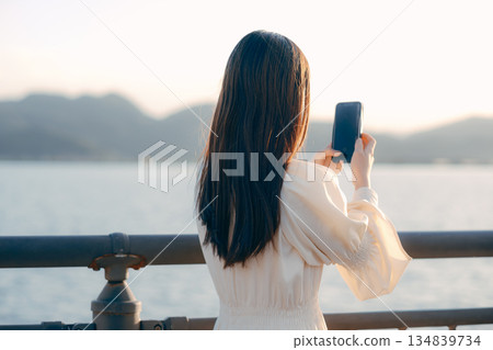 一位日本女子在河岸邊看著她的智慧型手機,背景是大海。 一位日本女子在河岸邊看著她的智慧型手機,背景是大海。 134839734