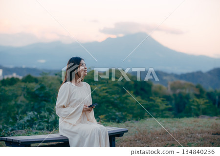 一位日本女子站在俯瞰城市景觀的山坡上,看著她的智慧型手機。 一位日本女子站在俯瞰城市景觀的山坡上,看著她的智慧型手機。 134840236