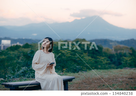 一位日本女子站在俯瞰城市景觀的山坡上,看著她的智慧型手機。 一位日本女子站在俯瞰城市景觀的山坡上,看著她的智慧型手機。 134840238