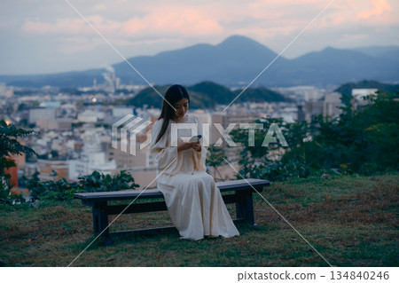 一位日本女子站在俯瞰城市景觀的山坡上，看著她的智慧型手機。 134840246