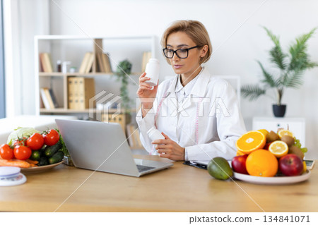 A female nutritionist consults with a client online, holding supplement bottles and surrounded by fresh fruits and vegetables. A female nutritionist consults with a client online, holding supplement bottles and surrounded by fresh fruits and vegetables. 134841071